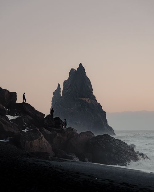 Free Silhouetted figures standing on rugged rocks by the sea at twilight, exploring breathtaking coastal scenery. Stock Photo
