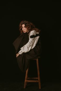 A young woman in retro fashion sits on a stool against a black background.
