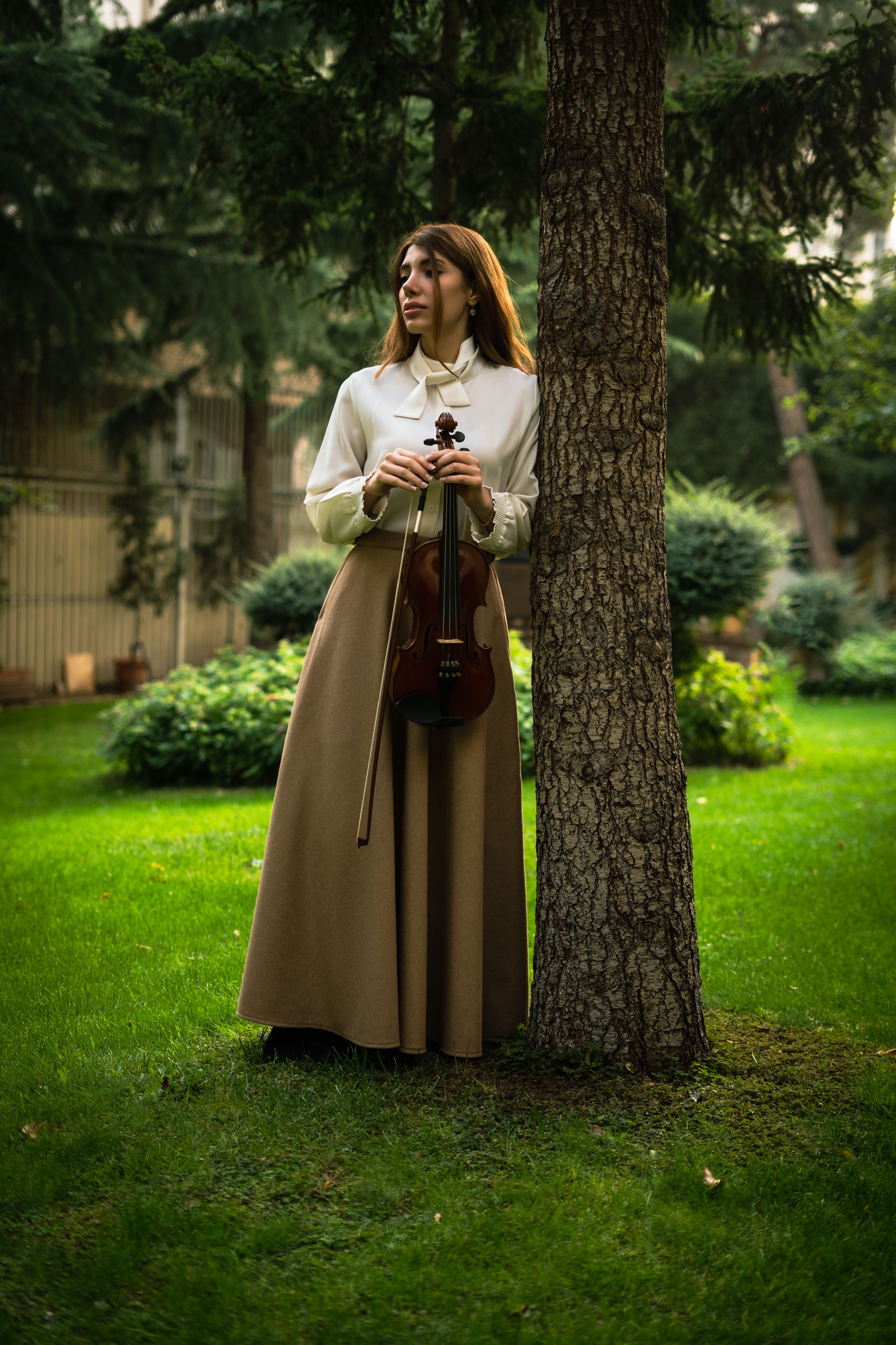 A woman stands with a violin in a peaceful Tehran garden, embracing nature and music.
