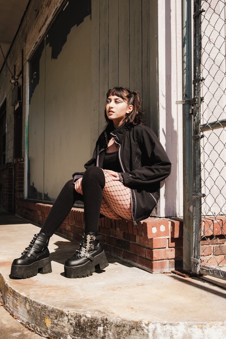 Black Goth Style Dressed Woman Sits On Brick Wall