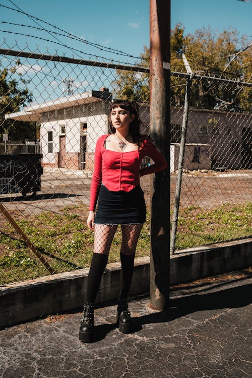 A woman in a red sweater and black stockings stands by a fence