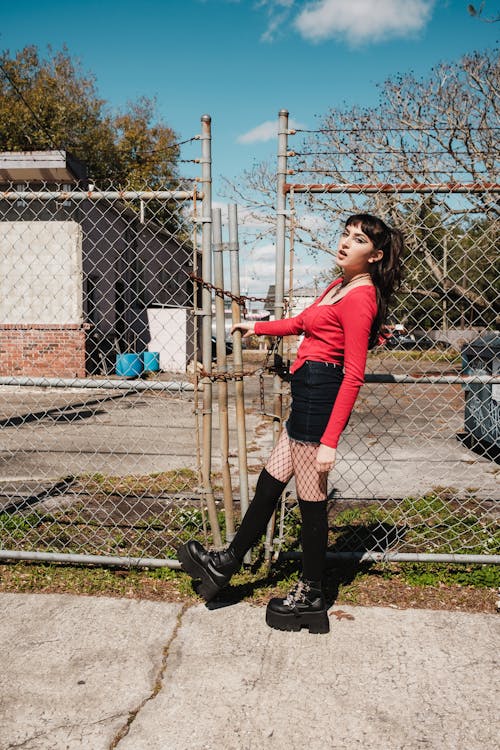 A woman in red top and black boots standing by a fence