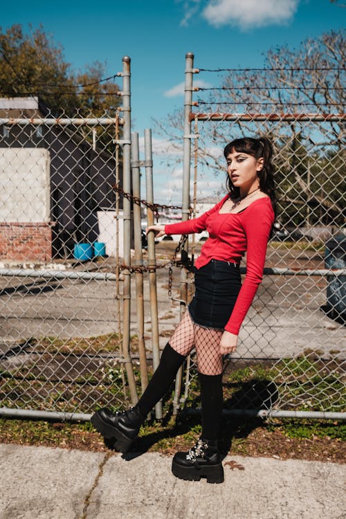 A woman in red shirt and black thigh high boots standing by a fence