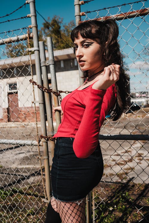 A woman in red top and black skirt posing near a fence