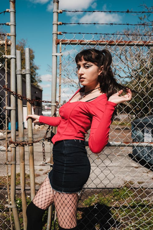 A woman in red shirt and black stockings posing near a fence