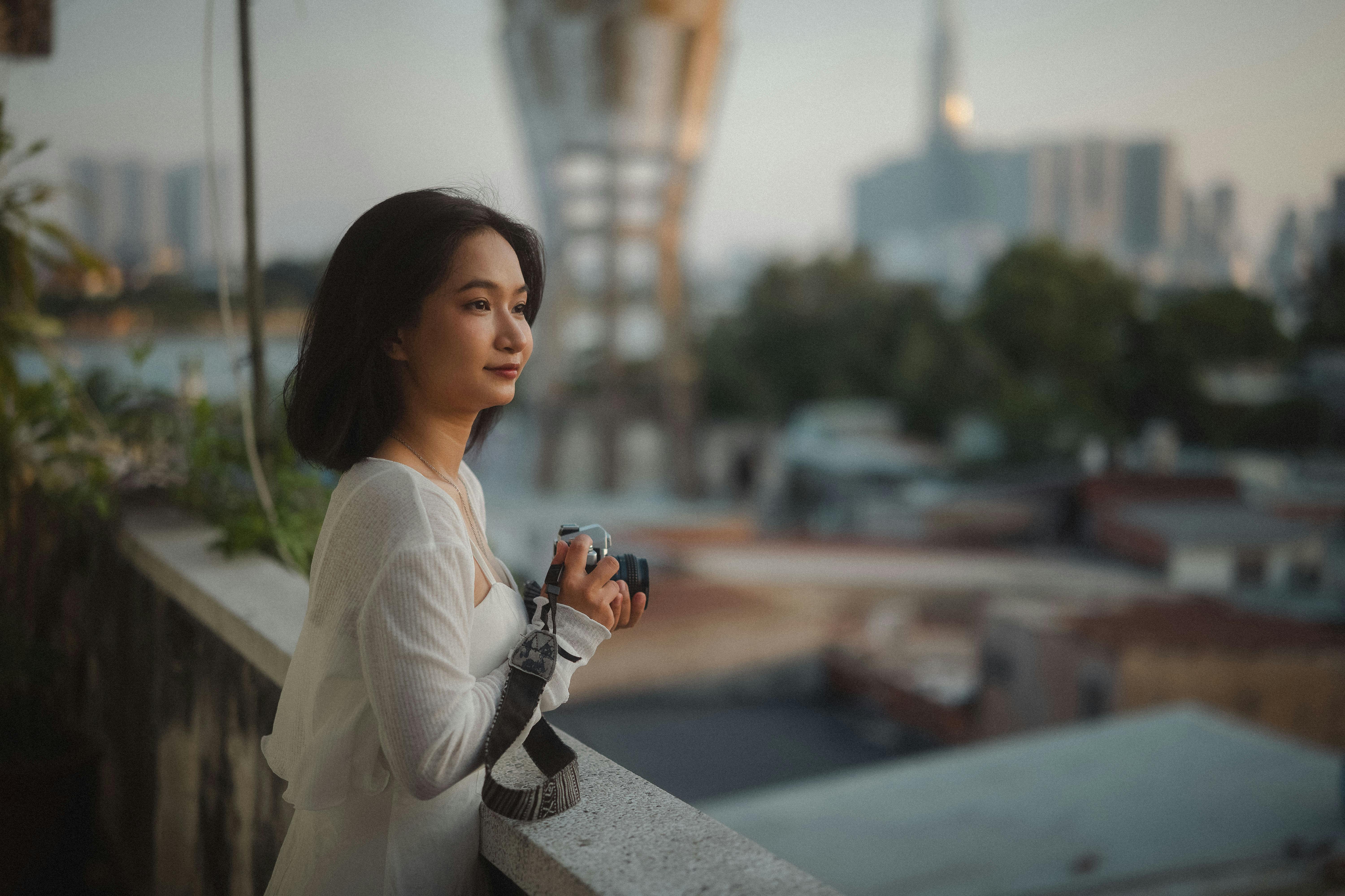 Woman capturing city skyline from a balcony at sunset, exuding calm contemplation.
