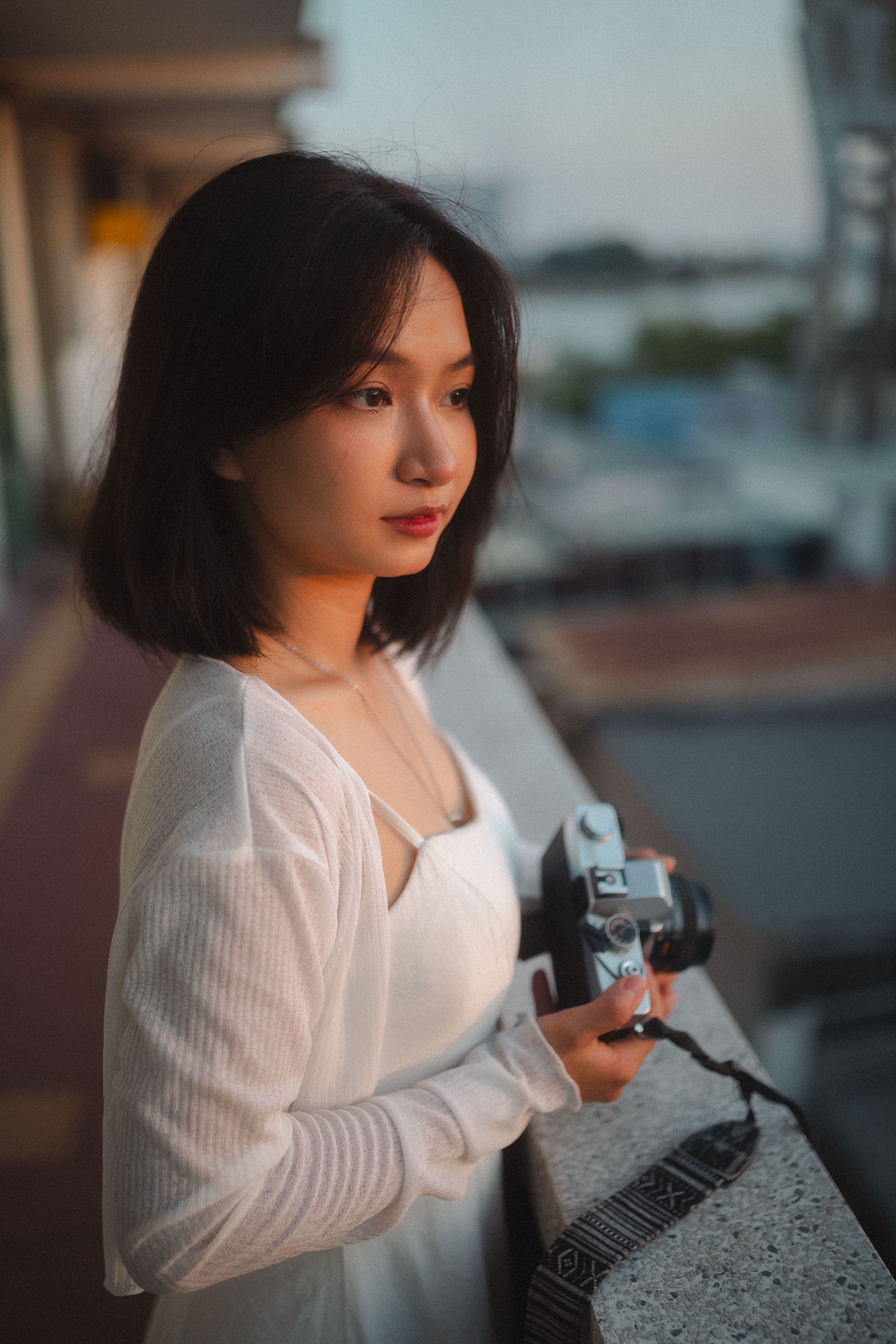Portrait of a young woman holding a camera, standing on a balcony during sunset.
