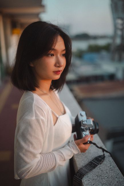 Portrait of a young woman holding a camera, standing on a balcony during sunset.