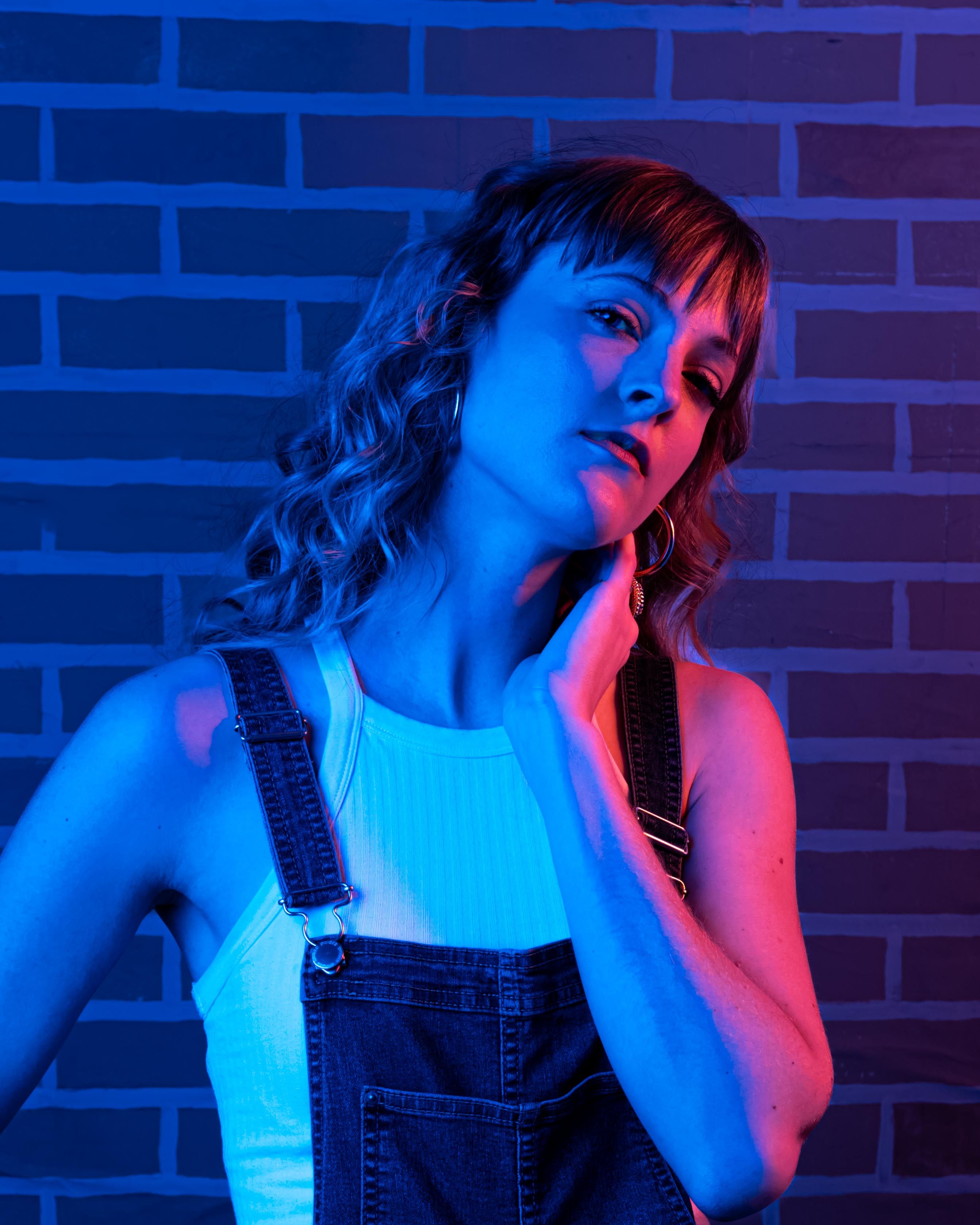 Portrait of a woman in denim overalls, highlighted by blue and red lighting against a brick wall.