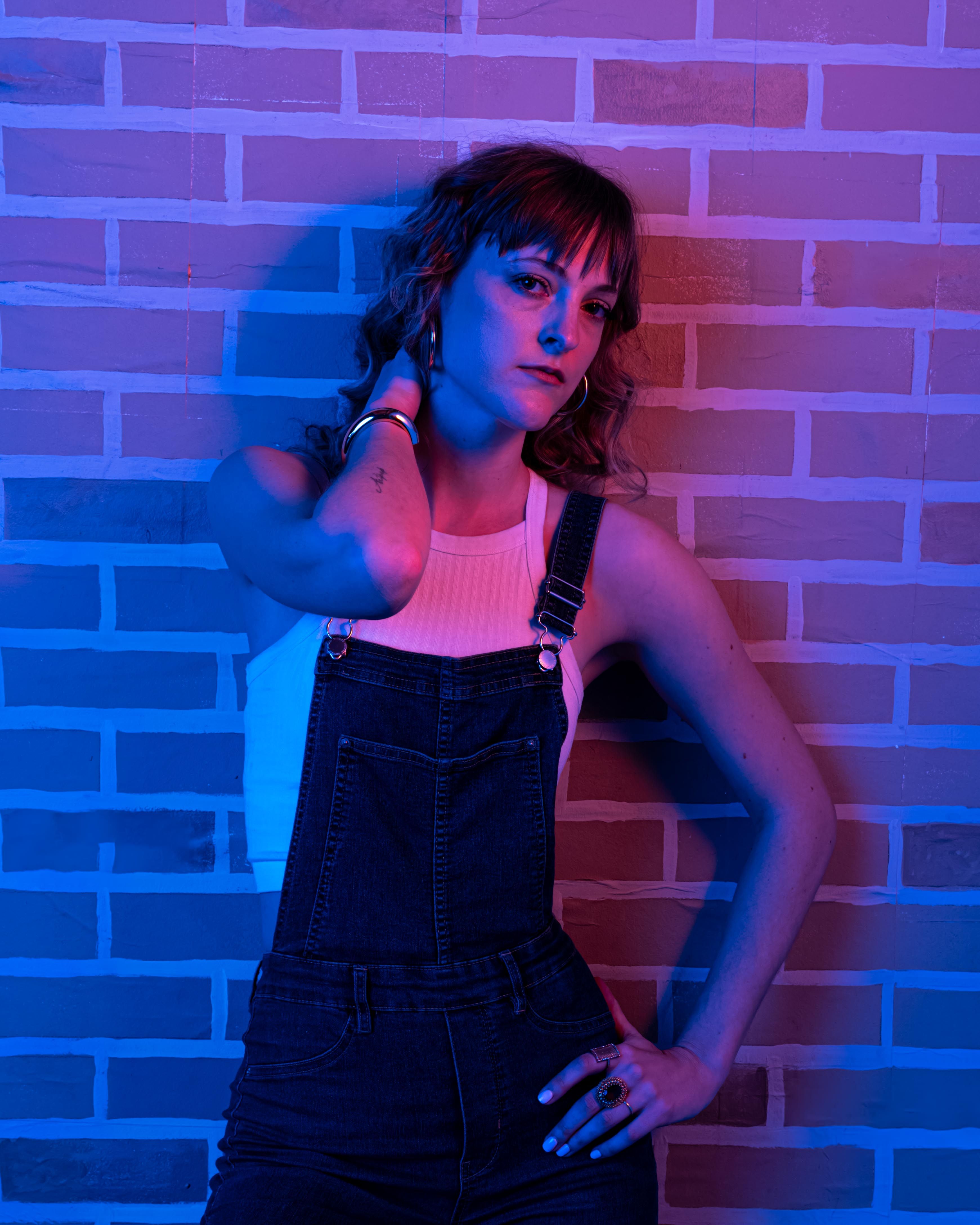 Fashionable woman in overalls posing against a brick wall with striking blue and red lighting.