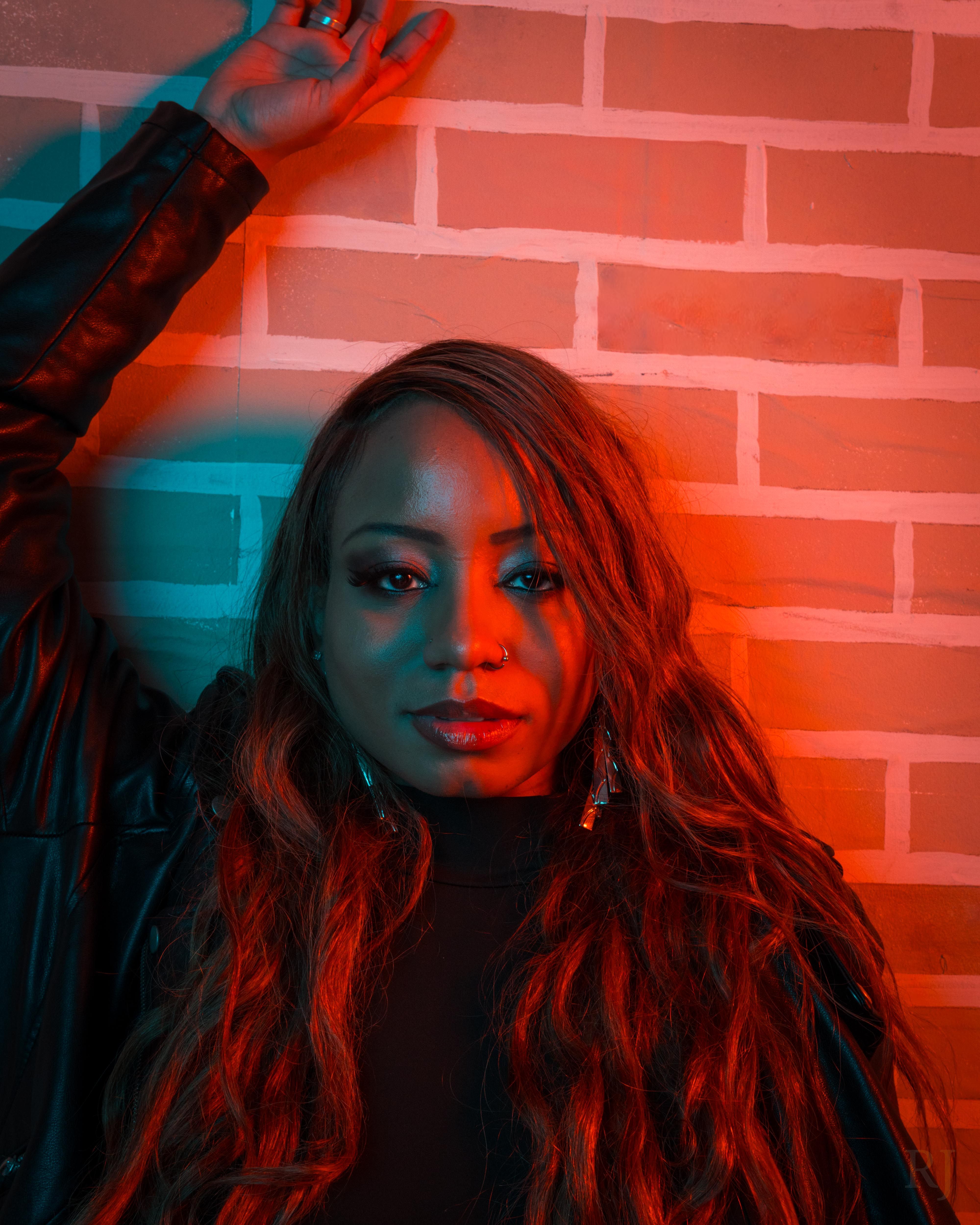 Portrait of a woman with dynamic red and blue lighting, standing against a brick wall.