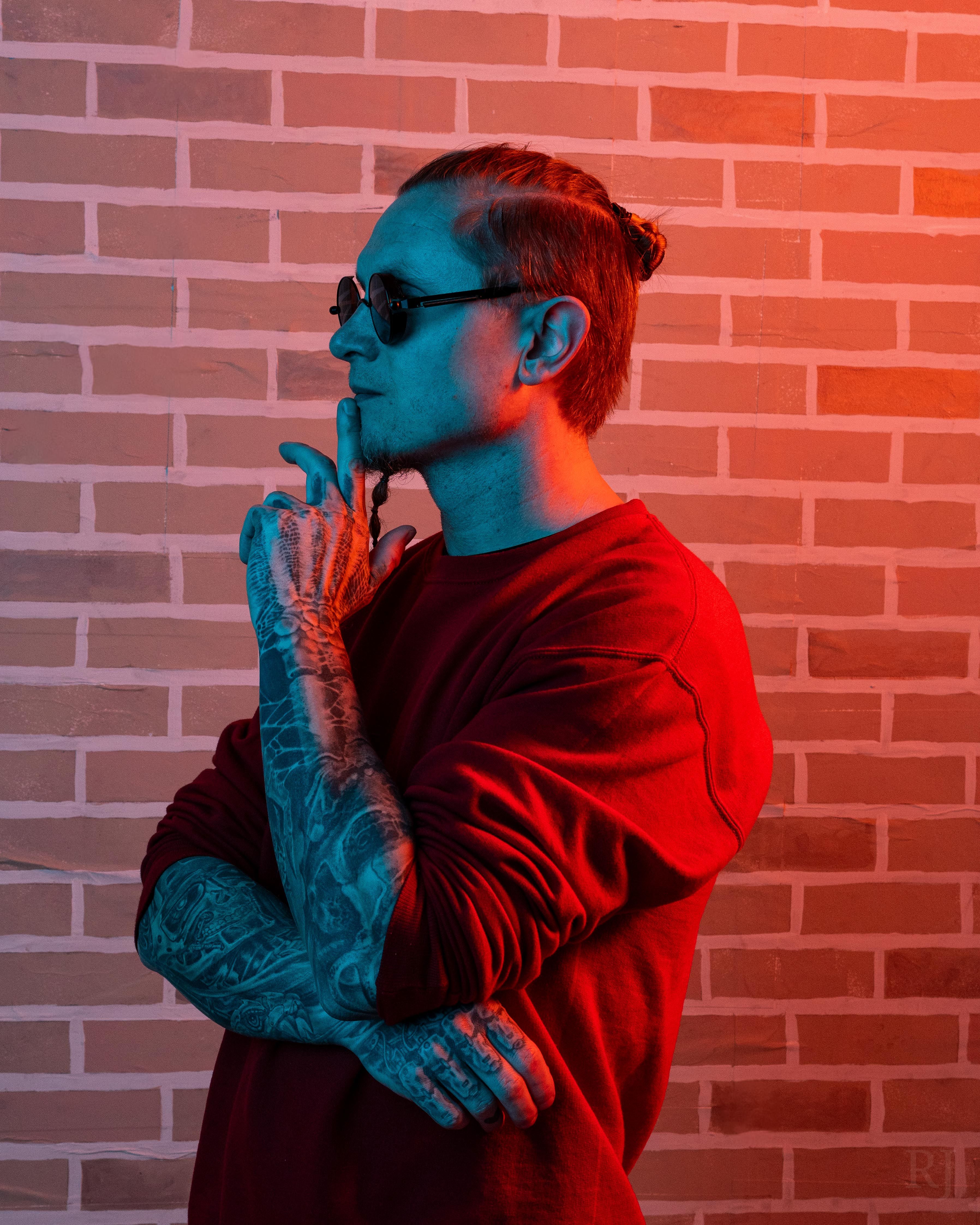 Tattooed man in sunglasses and red shirt poses thoughtfully against a brick wall with dramatic lighting.