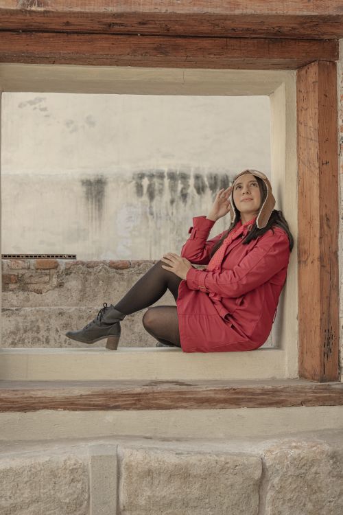 Free A woman in a red jacket and aviator hat sits in a window frame looking up thoughtfully. Stock Photo