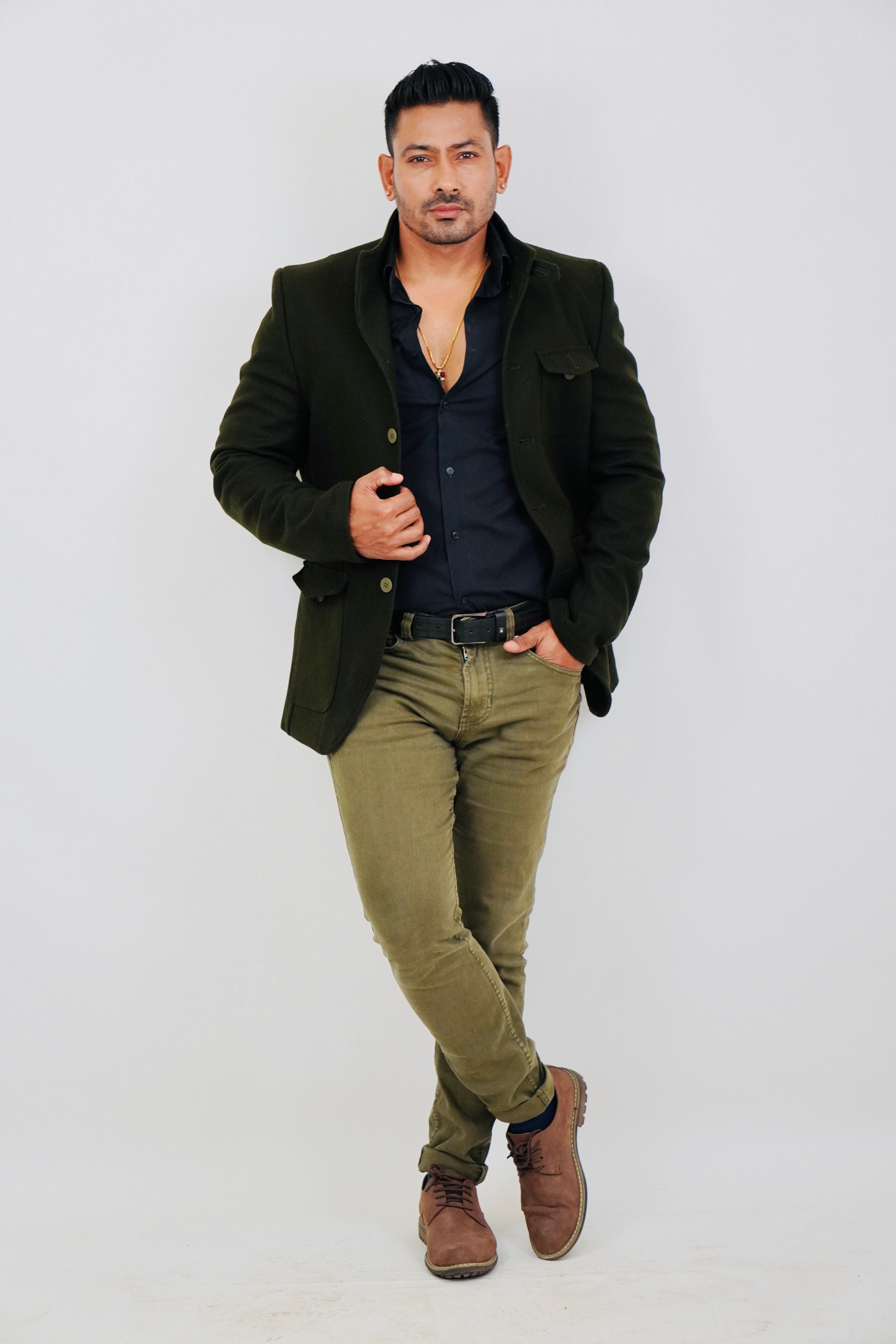 Confident male model poses stylishly in a dark green jacket and brown shoes against a white background.