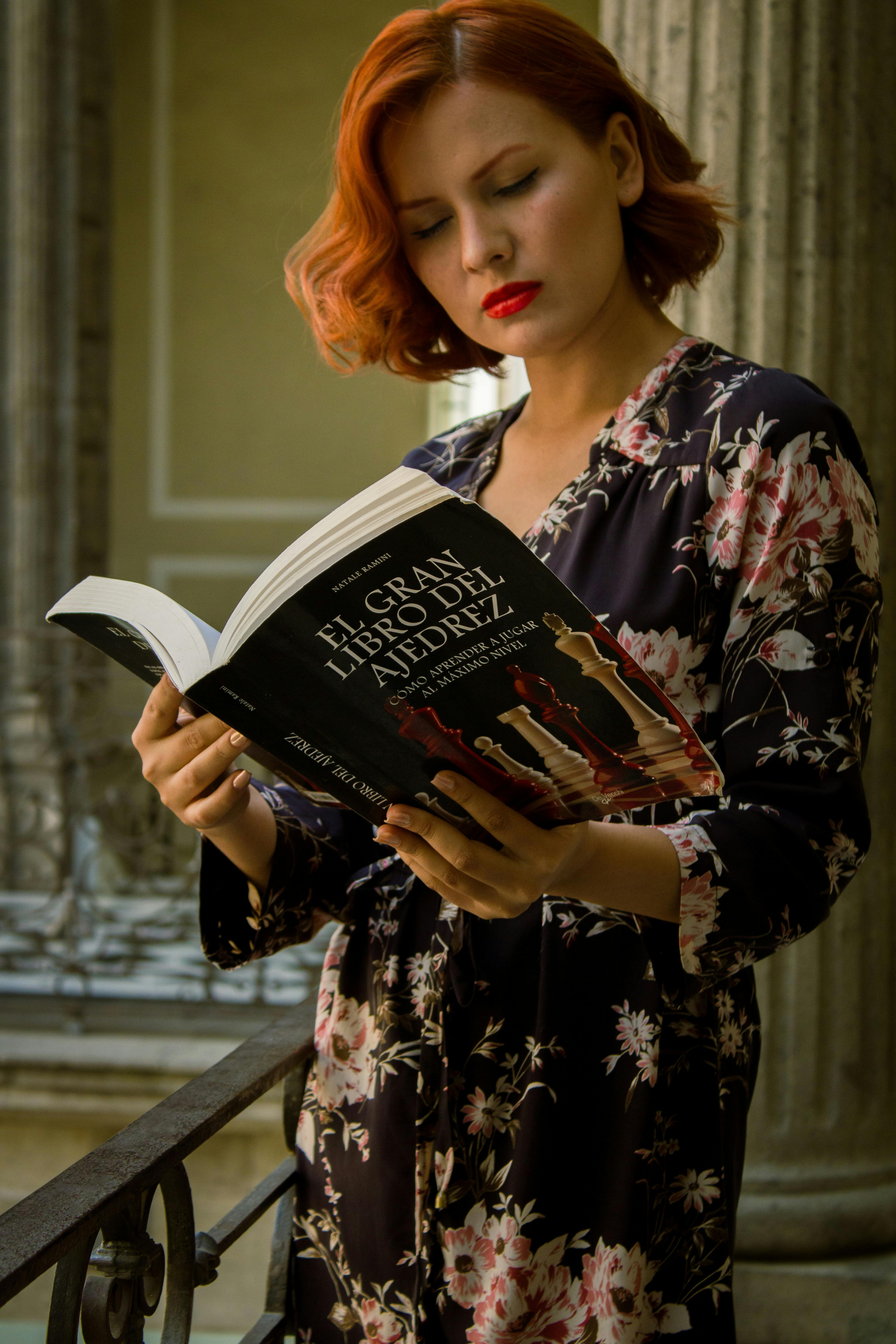 Redhead woman in floral dress reading a chess book on a balcony.