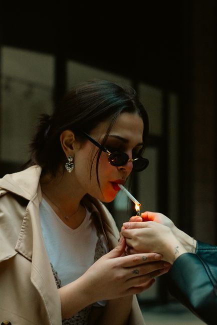 A young woman lights a cigarette with a lighter while wearing sunglasses, set against an urban backdrop.