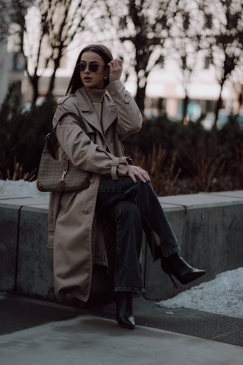 A woman in a trench coat and sunglasses sitting on a bench