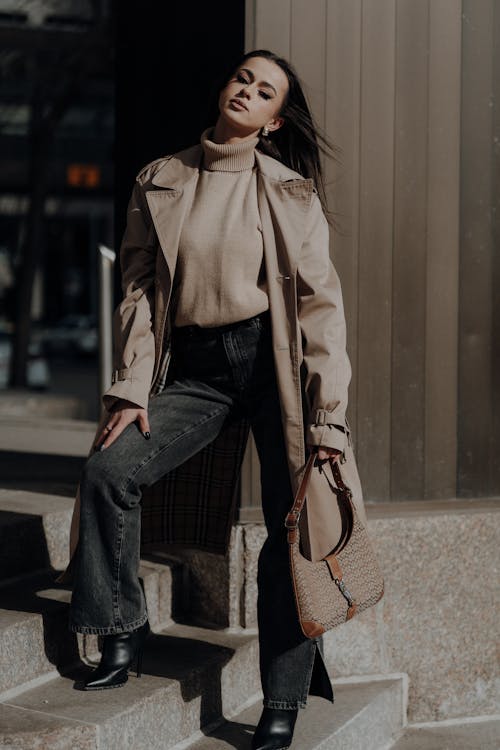 A woman in a trench coat and jeans is posing on steps