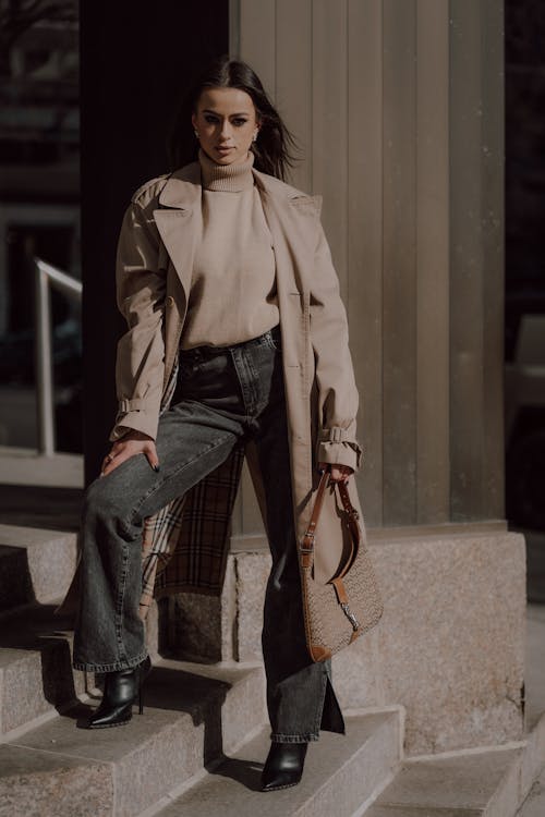 A woman in a trench coat and jeans is standing on steps