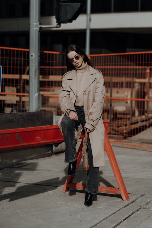 A woman in a trench coat and black jeans sits on a bench