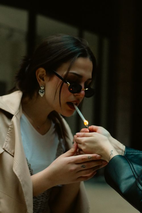 Free Portrait of a stylish woman in sunglasses being assisted to light her cigarette in a casual setting. Stock Photo