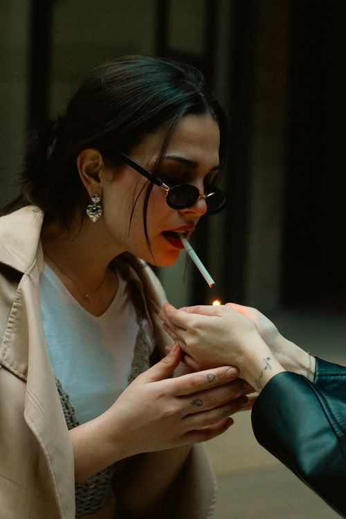 Free Stylish woman in sunglasses lighting a cigarette outdoors in soft lighting. Stock Photo