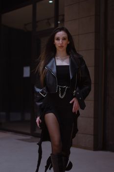 Stylish woman in black dress and leather jacket confidently poses on urban street.
