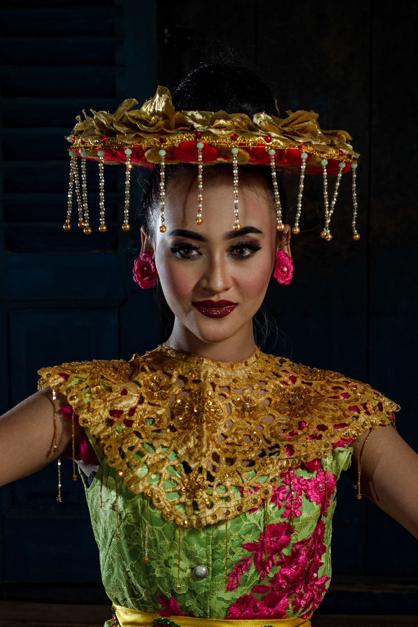 Portrait of an Asian woman in traditional Indonesian attire with ornate hat and floral details.