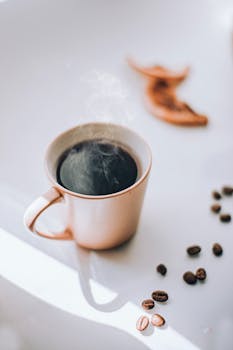 Elegant photo of a steaming coffee cup surrounded by scattered beans on a bright surface.