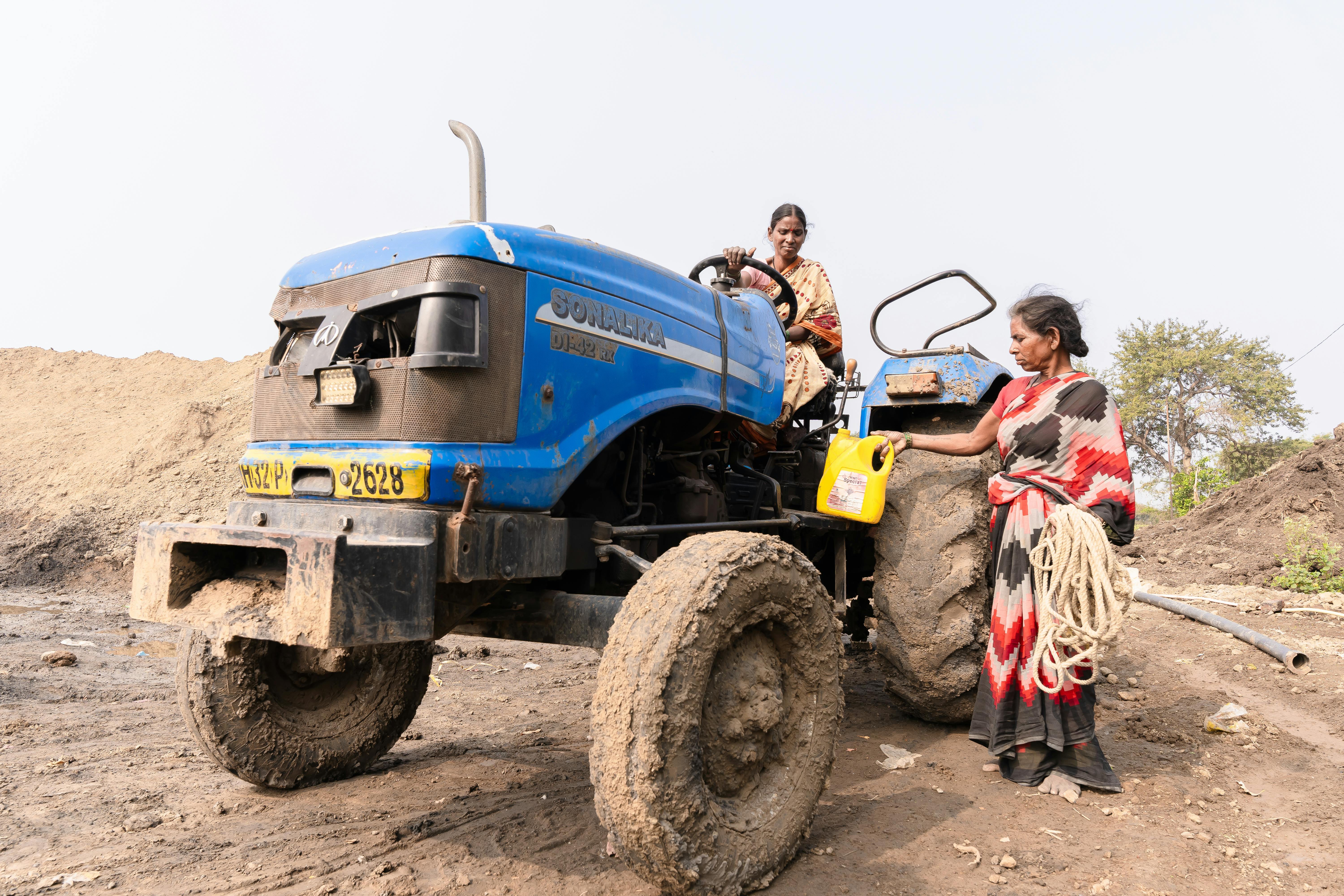 Two Indian women farmers working near a muddy blue tractor on a countryside farm.