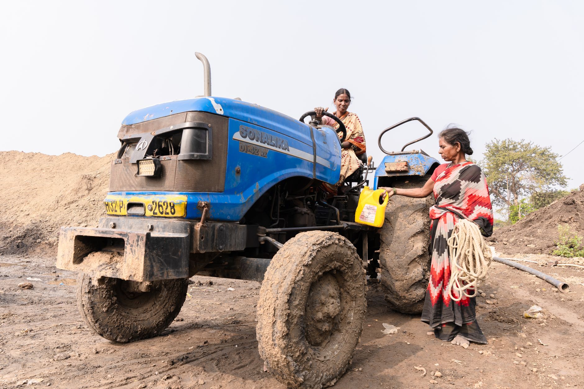 Two Indian women farmers working near a muddy blue tractor on a countryside farm.