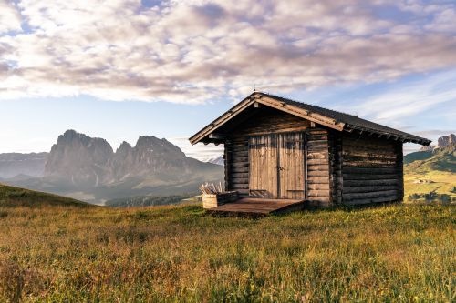 Free A serene wooden shed amidst lush grasslands with a mountain backdrop under a bright sky. Stock Photo