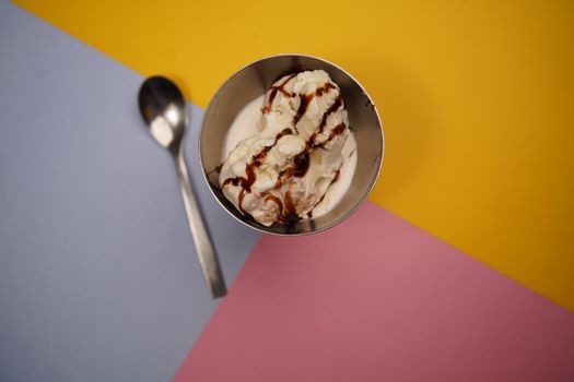 Delicious ice cream in a metal bowl with chocolate drizzle on a vibrant geometric background.