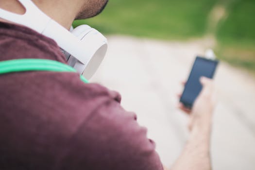 Adult man holding a smartphone outdoors with headphones around the neck, blurred background.