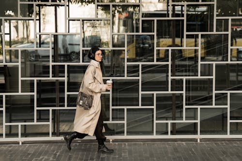 Young Woman in a Beige Coat and Headphones Hurrying Down the Sidewalk with a Cup of Coffee