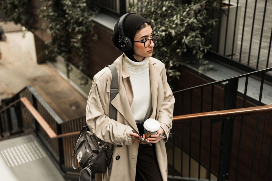 Young woman in a beige coat and hijab walking down stairs with coffee and headphones.