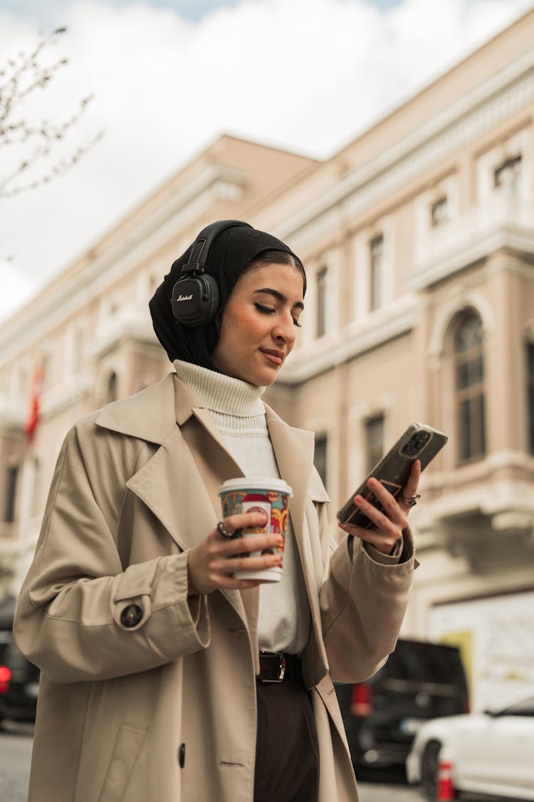 Woman Wearing A Beige Coat And Headphones, Walking In A Street With A Coffee And Phone