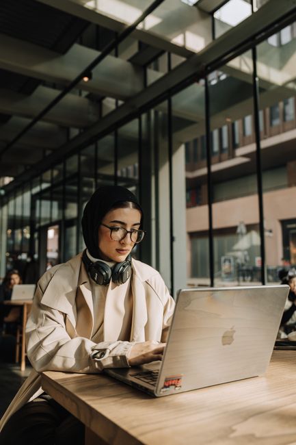 Focused Muslim woman working on a laptop in a sunlit coworking space.