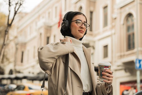Young Woman Wearing a Coat and Headphones, Walking in a Street with a Coffee