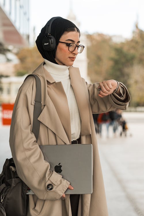 Photo of a Young Woman Wearing Headphones and a Beige Coat, Holding a Laptop on a Street