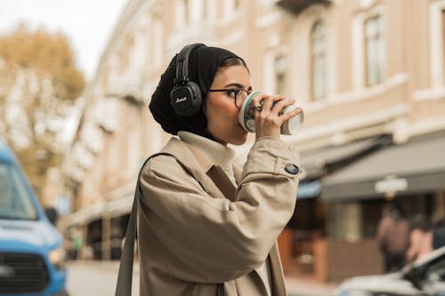 Woman Wearing a Headscarf and Headphones, Drinking Coffee on a Street