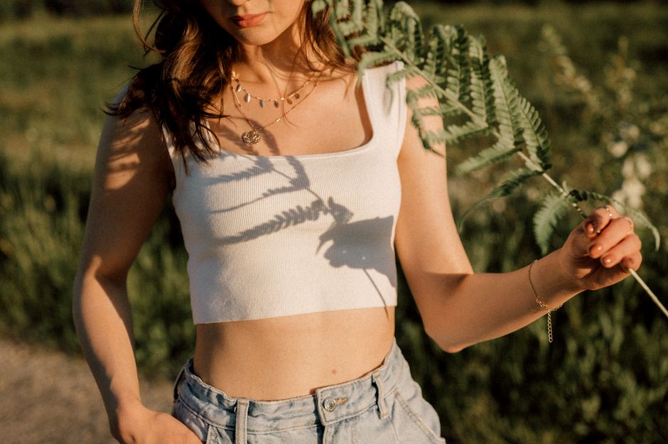 Young woman holding fern leaves in a sunlit field, casting shadows on her top.