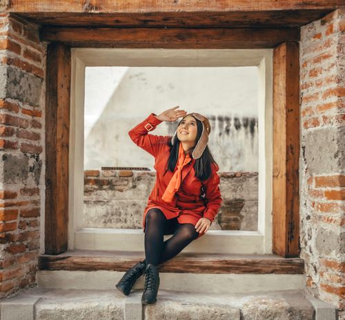 Free Stylish woman in red jacket with aviator hat sitting in a rustic window frame, looking up thoughtfully. Stock Photo