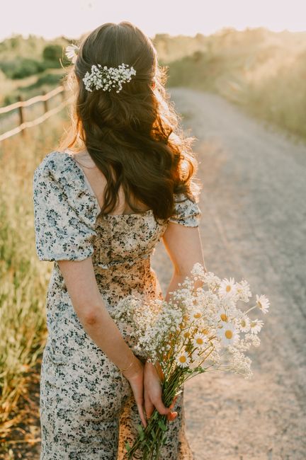 A woman holding a bouquet of daisies on a sunlit path, wearing a floral dress.