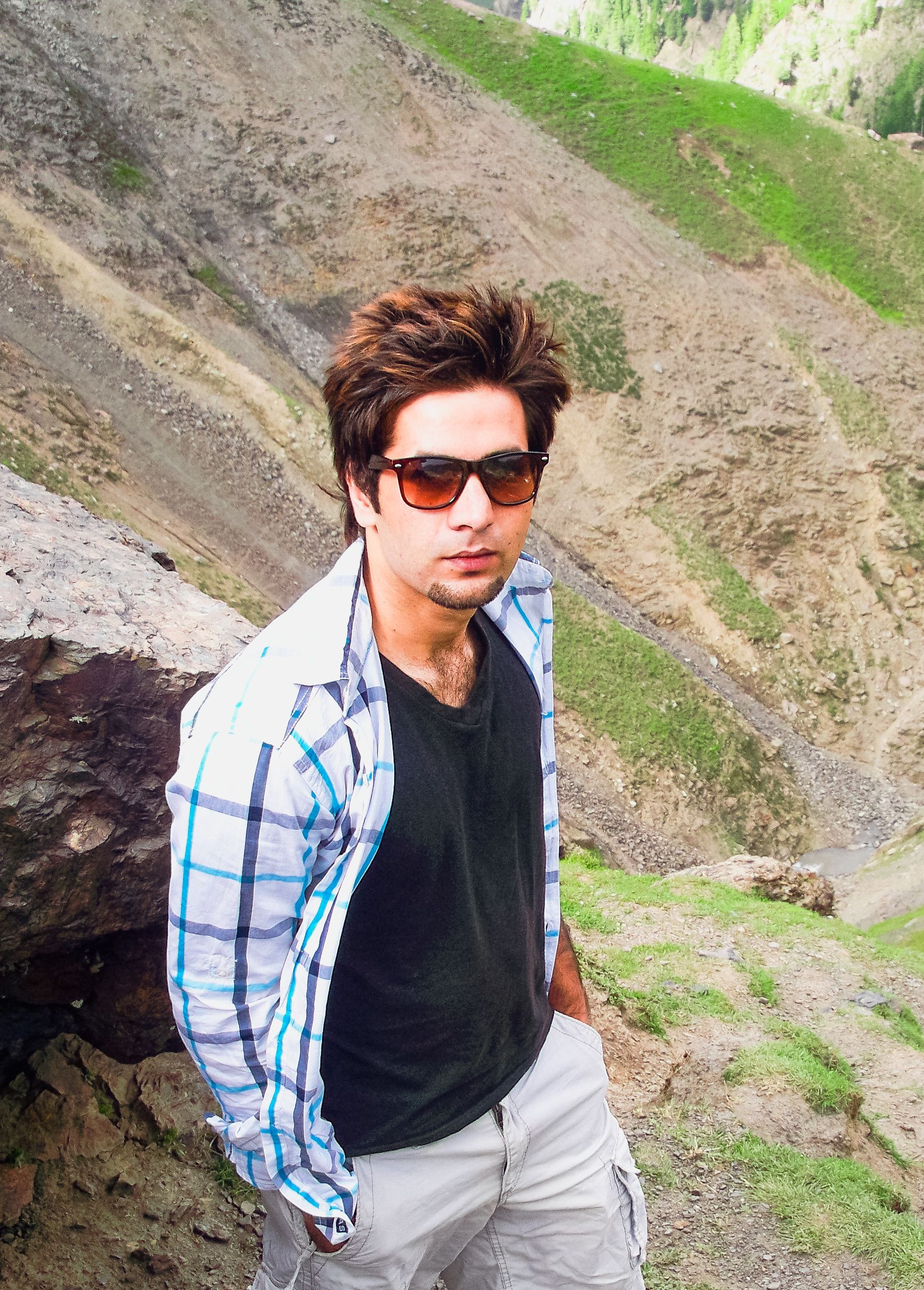 Man in casual attire with sunglasses stands confidently in a rugged outdoor setting. Bright day.