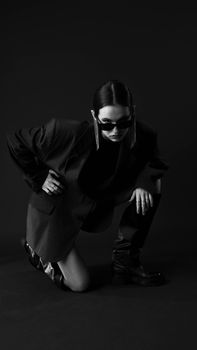 Stylish woman in sunglasses crouching in black suit in a dramatic black and white studio shot.