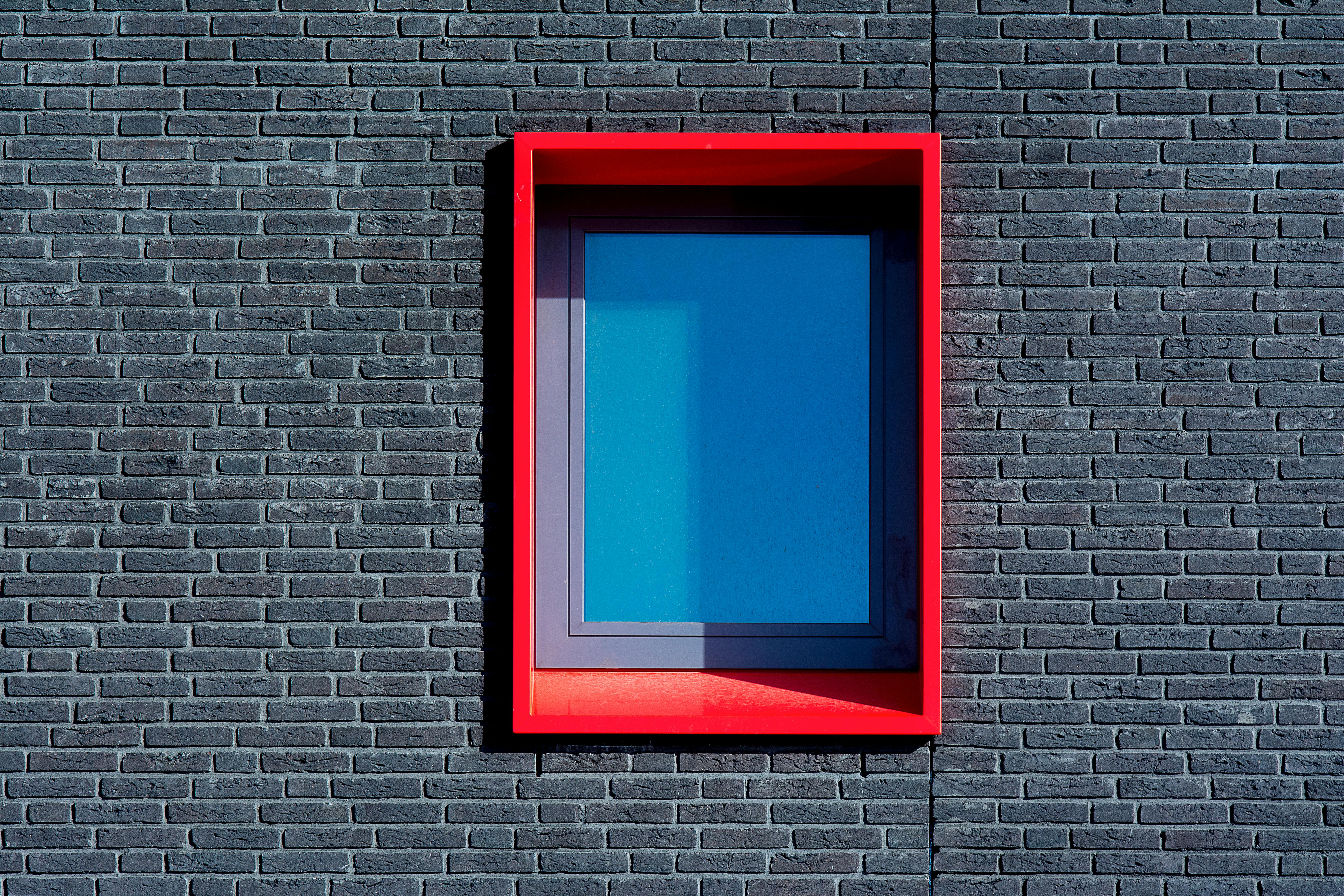 A minimalist urban brick wall featuring a bright red window frame with a blue pane.