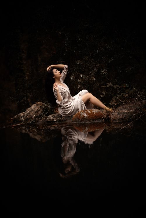 Free A woman in a white dress poses gracefully by a tranquil riverbank, reflecting in the water. Stock Photo