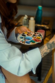 Woman with tattooed arm holding colorful paint palette, capturing creative artistry indoors.