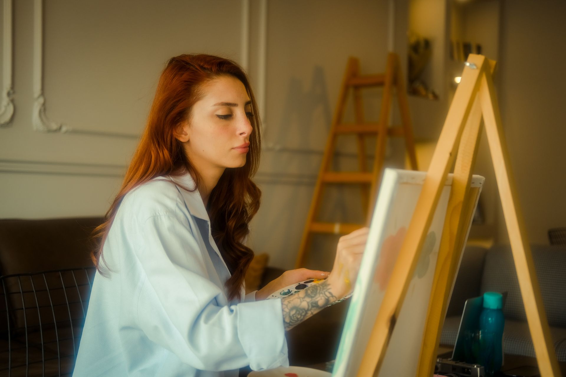 A focused young woman artist with red hair painting on a canvas in an art studio.